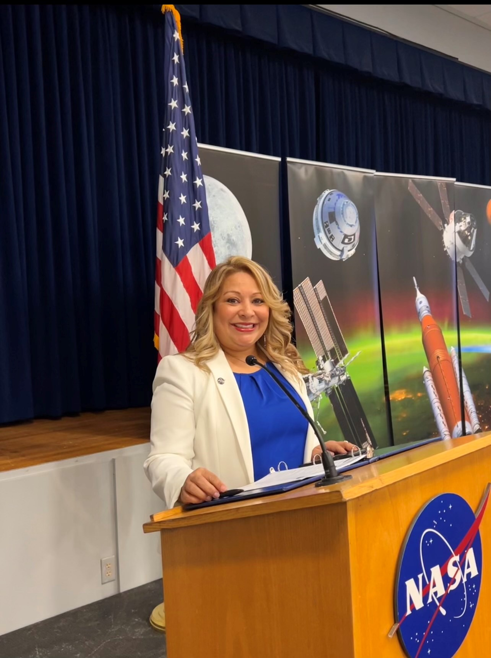 A person stands in front of a podium with a blue shirt and white suit jacket on and an American flag behind them.